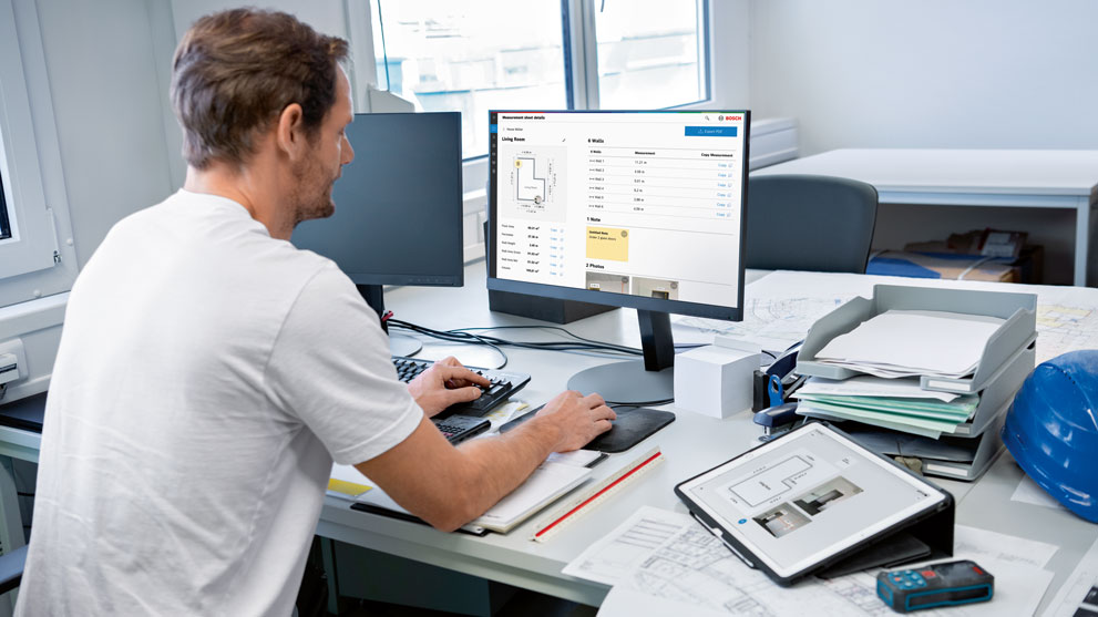 A man is working on a computer in a bright office with construction plans.