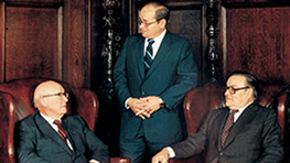 Three men in suits are sitting and standing in an elegant room.