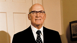 An older man in a suit with a tie and glasses stands in front of a wall.