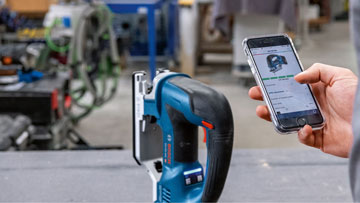 Bosch Professional cordless jigsaw is on a table, one hand is holding a phone.