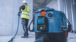 A blue Bosch Professional industrial vacuum cleaner stands in the foreground, while a worker in a yellow vest and helmet works behind it.