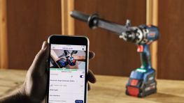 A person holds a smartphone in their hand, showing a Bosch Professional battery screw. The screw is on a wooden surface. The tools are red and blue, and the environment looks bright and modern.