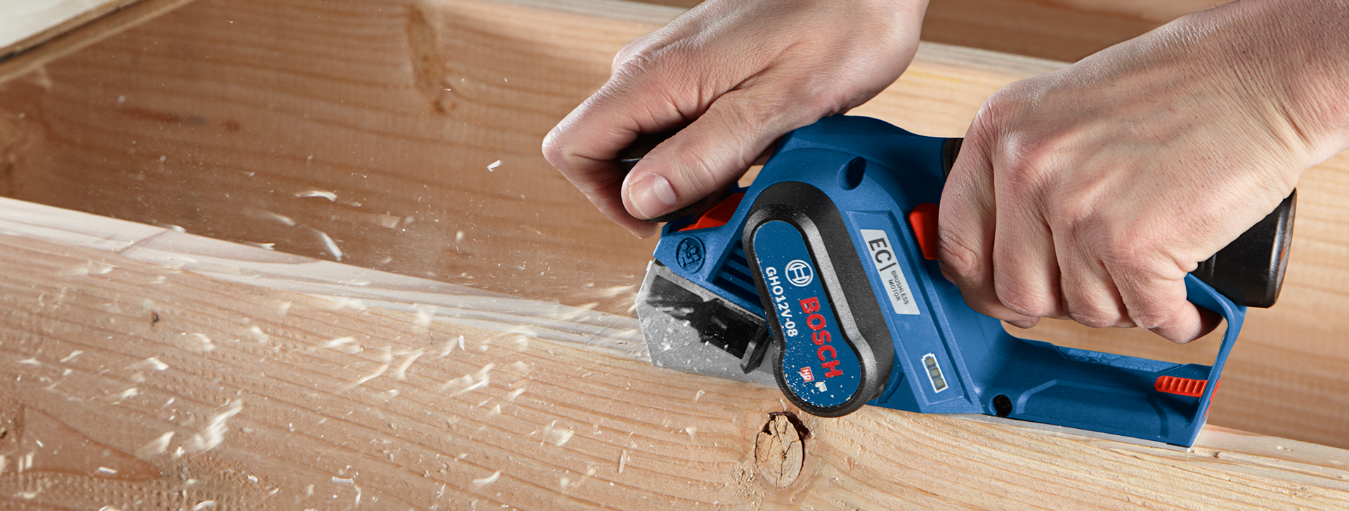 A person is using a blue Bosch planer while wood shavings fly from a wood block.