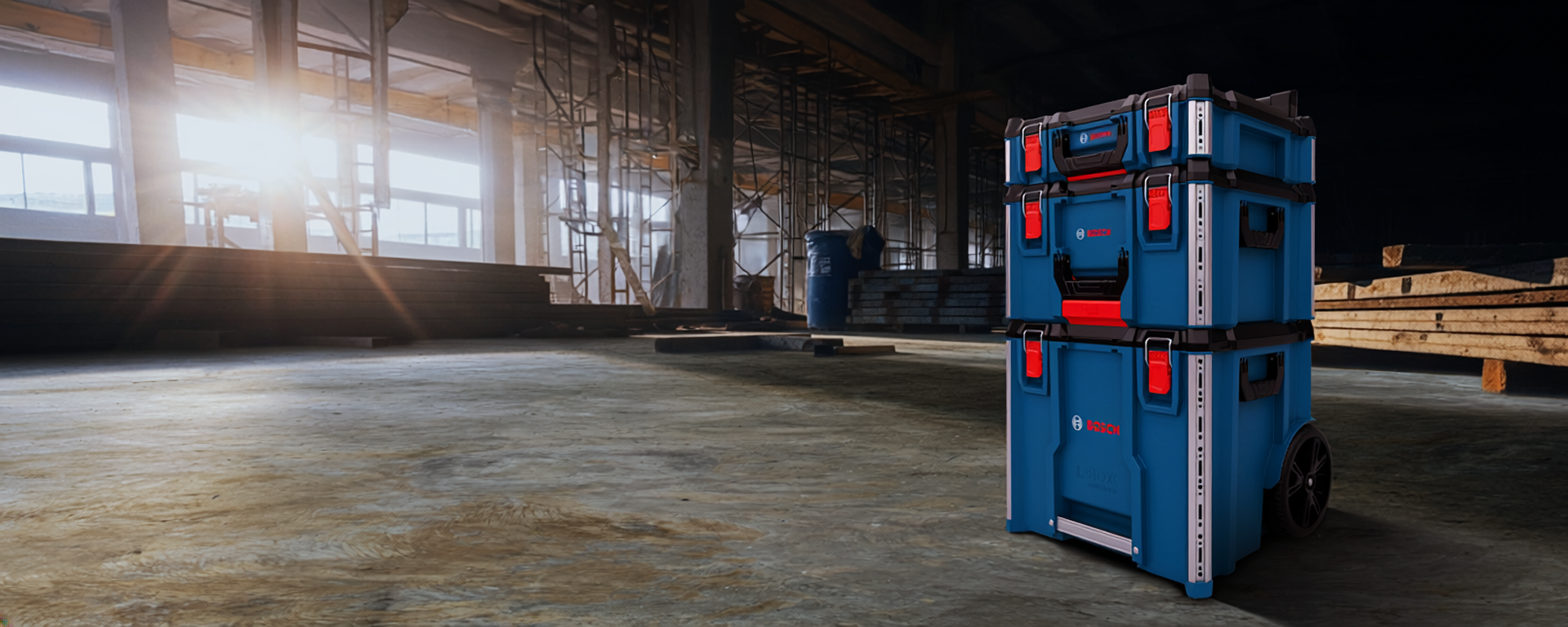 A blue Bosch Professional tool case stands in the foreground on a concrete floor at a construction site. In the background, wood piles and steel frameworks are visible, while sunlight streams through large windows.