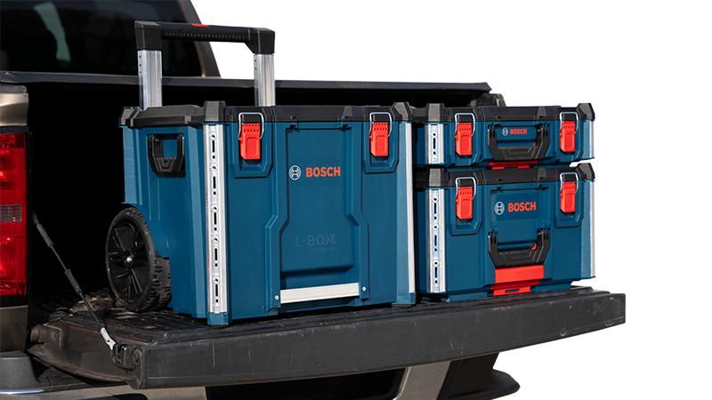 Two large blue toolboxes from Bosch are on the bed of a pickup truck. The boxes have orange latches and are on wheels with a retractable handle. The background shows the truck in a bright, sunny environment.