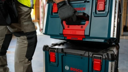 Un travailleur en gilet de sécurité jaune soulève une boîte à outils Bosch Professional verte avec des accents rouges. Les boîtes sont sur un sol gris, partiellement visible.