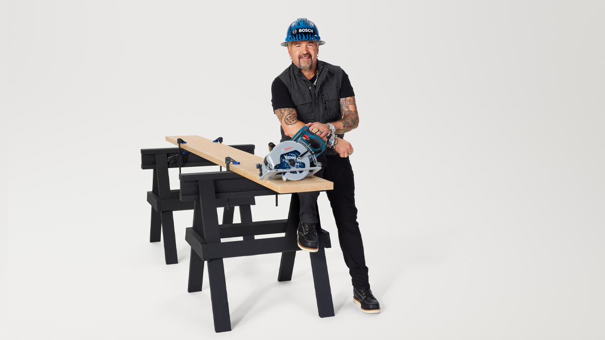 A man with tattoos stands at a table, holding a circular saw and smiling. He is wearing a blue helmet.