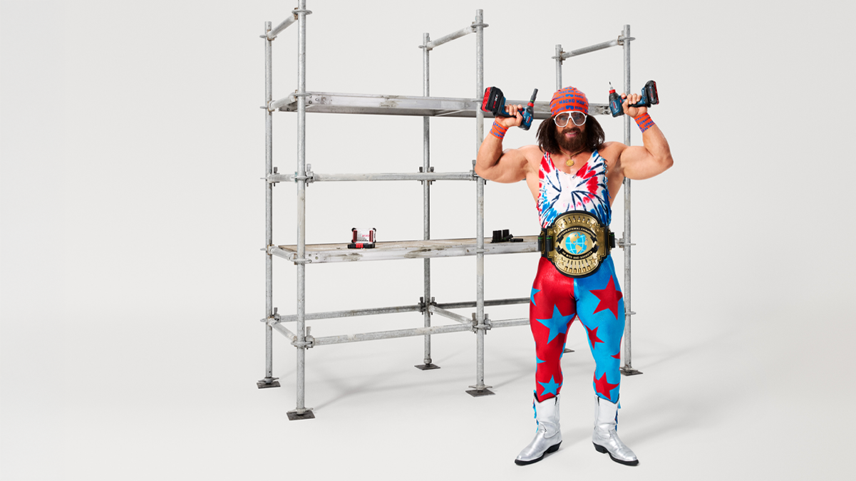 A muscular man in a colorful suit with stars holds two power drills overhead, in front of a scaffold.