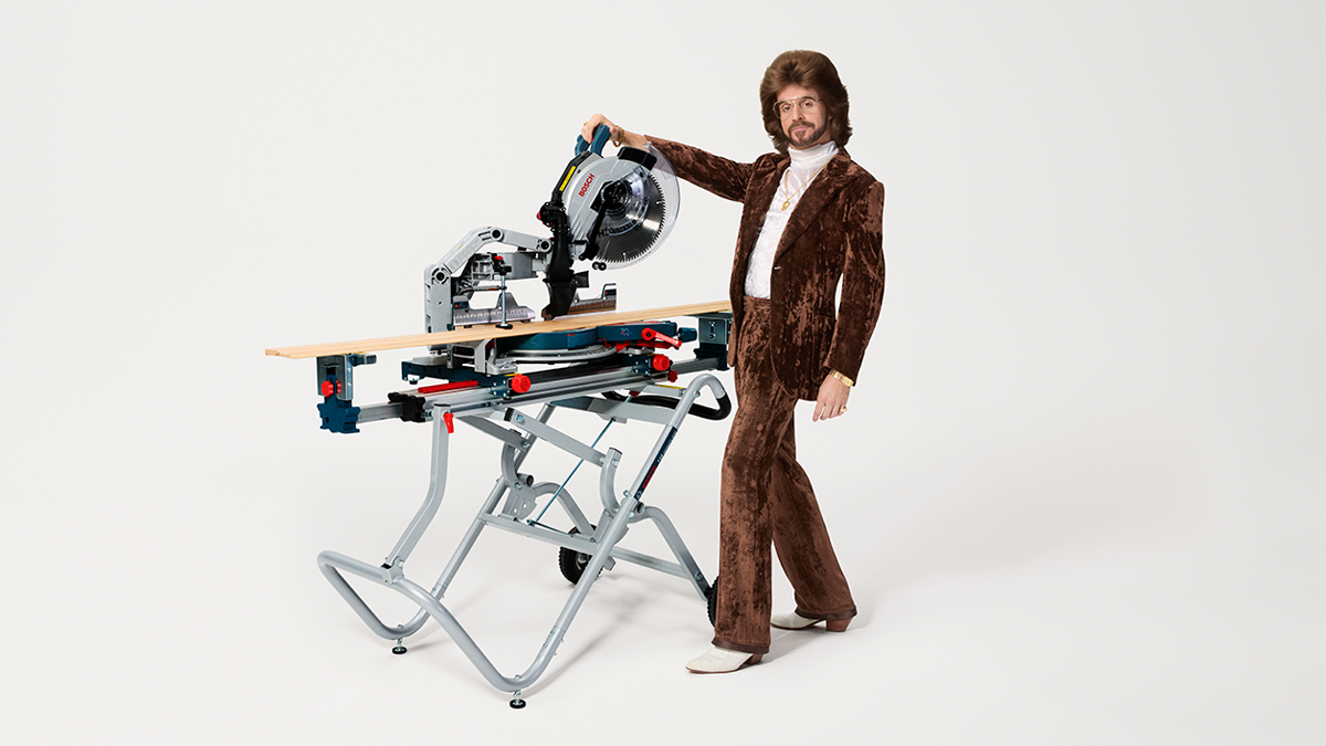 A man in a brown velvet suit stands next to a table saw mounted on a table. He has a striking hairstyle and wears a gold chain.
