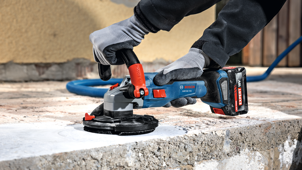 A person in gloves holds an electric sander over a gray concrete floor. A wall is visible in the background.