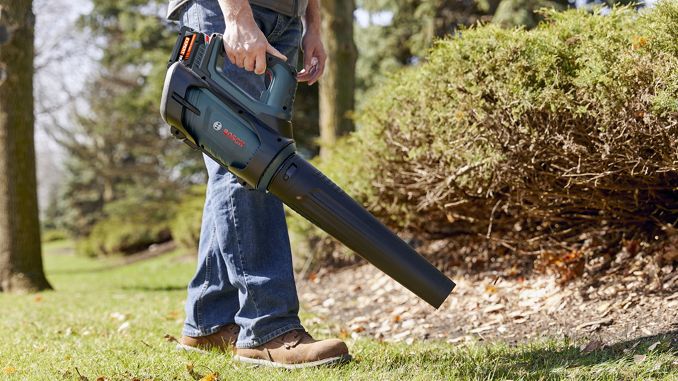 A man in jeans and brown shoes holds a leaf blower. He stands on green grass next to a bush.