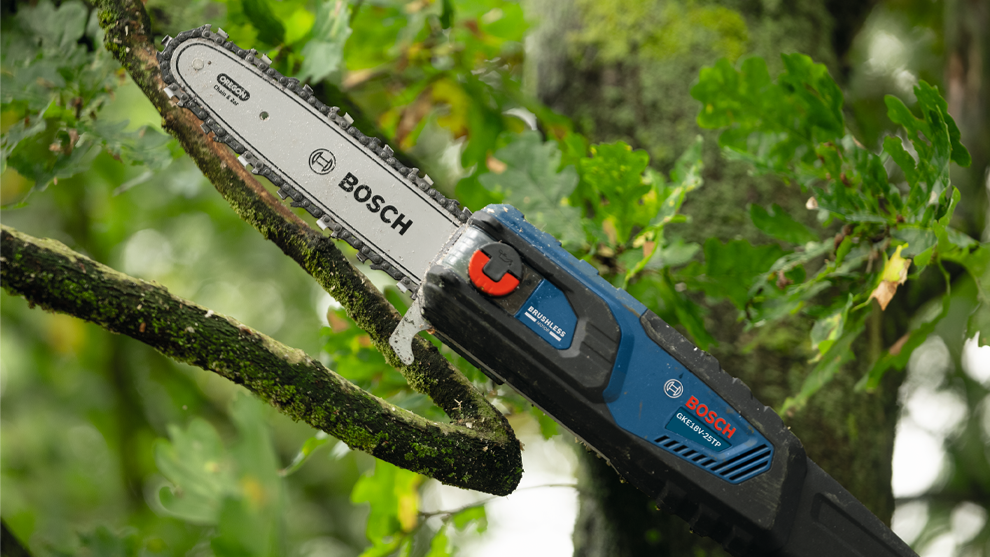 An electric chainsaw from Bosch is cutting a thick branch on a green tree.