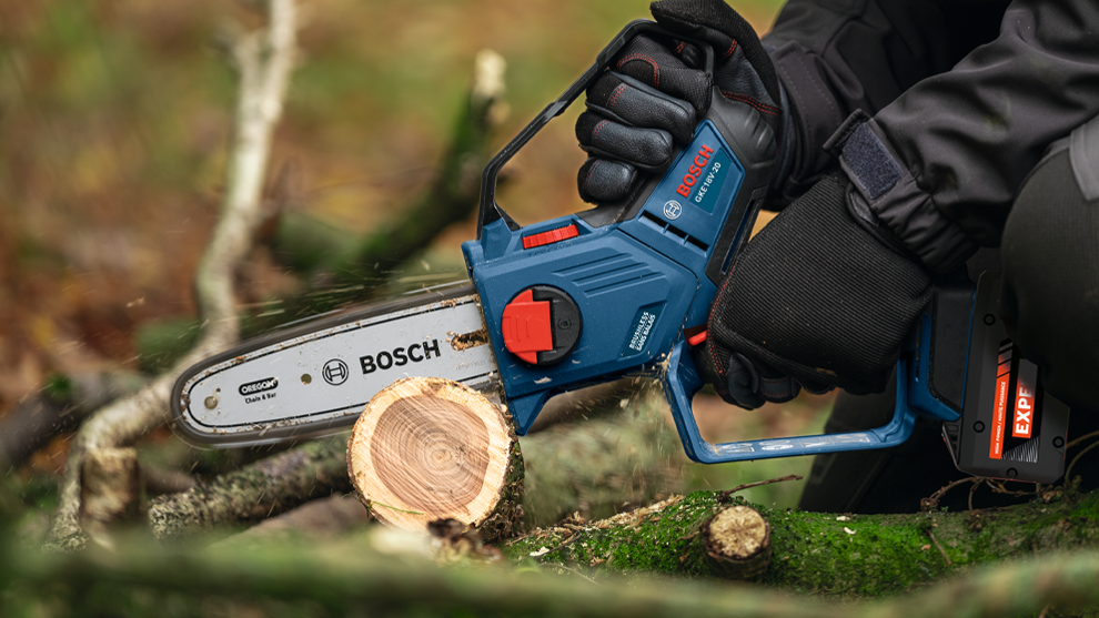 A person in black clothing holds an electric chainsaw cutting wood. In the background, branches and a forest are visible.