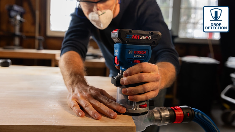 A man in a blue sweater and protective mask is using a Bosch Professional router on a wooden table. The machine is blue with red accents and has a vacuum cleaner connection. In the background, tools and a window front are visible, allowing daylight to enter.