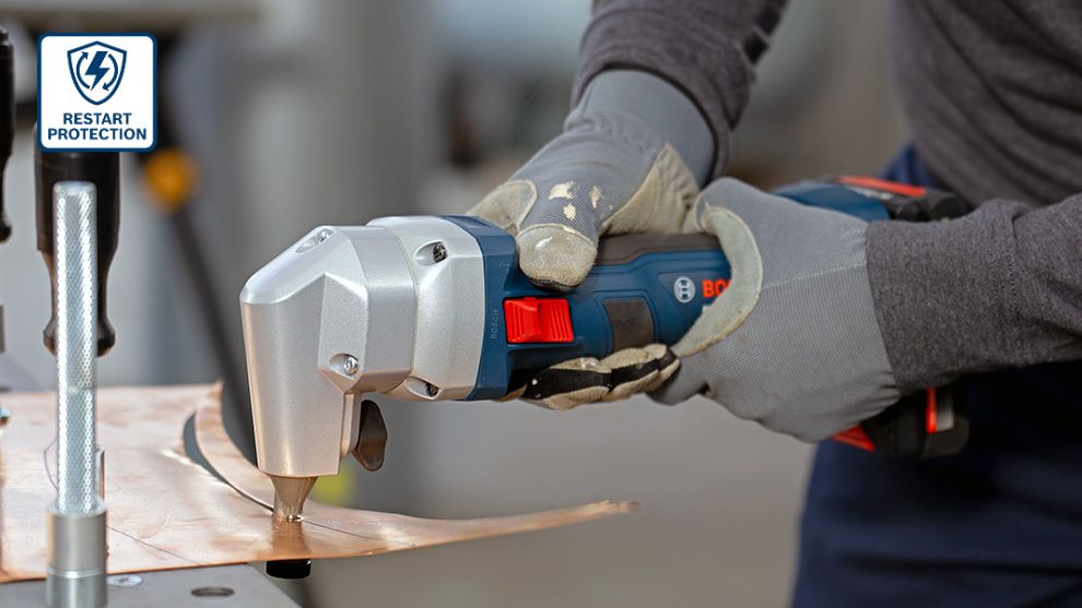 A Bosch Professional battery cutter in blue and gray is cutting copper sheet. The user is wearing gray gloves.