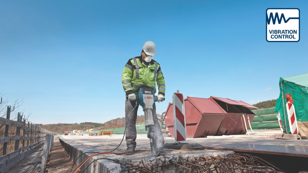 A worker in a neon yellow jacket and helmet operates a Bosch Professional demolition hammer at a construction site. In the background, green construction tarps and machinery are visible.