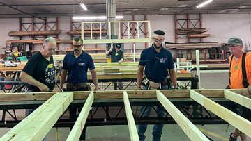 Four craftsmen are working on wooden beams in a workshop.