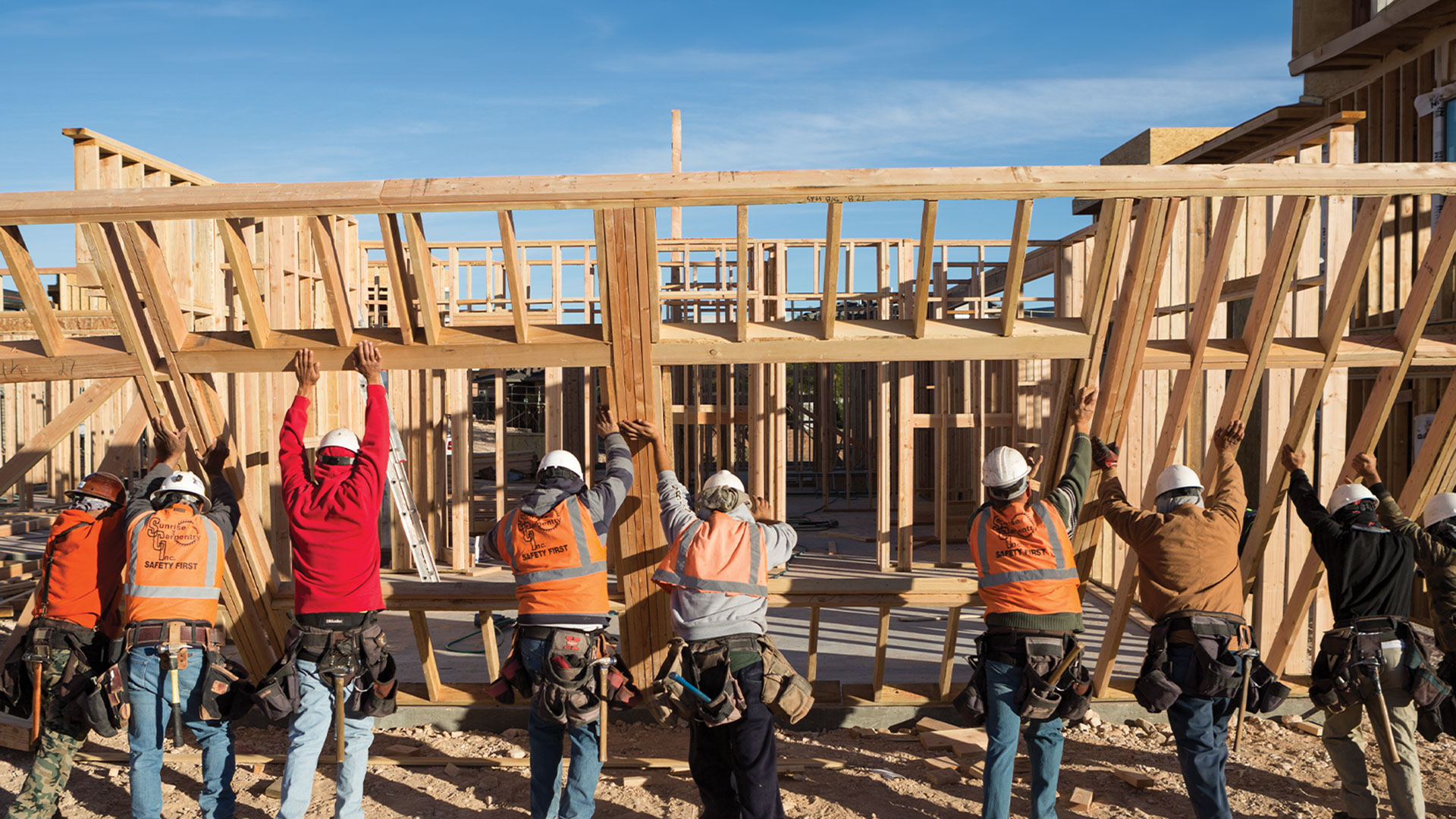 Five construction workers are lifting a wooden beam in a scaffold.