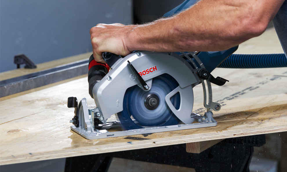 A hand holds a Bosch electric circular saw, working on a wooden board. Chips are flying.