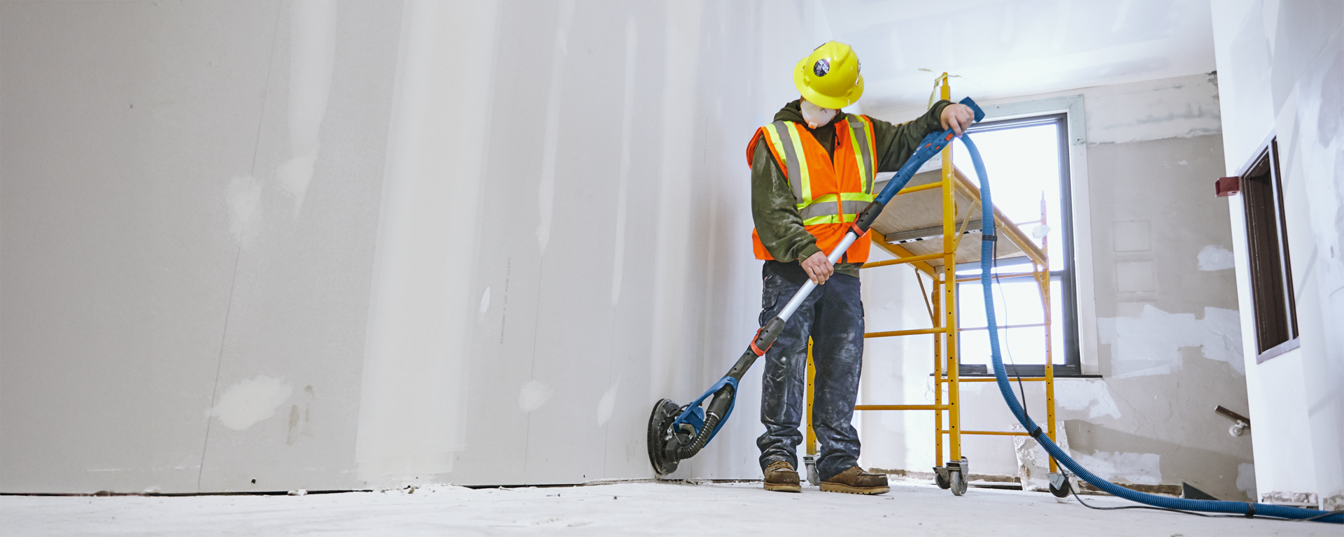 A worker wearing a hard hat and vest is using a vacuum cleaner in a room.