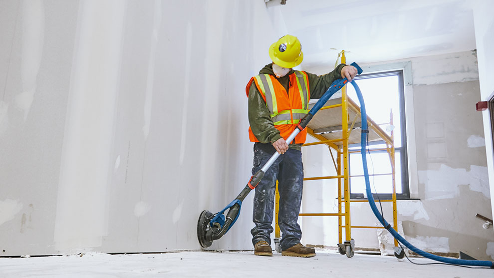 A construction worker wearing a helmet and safety vest is using a vacuum cleaner.